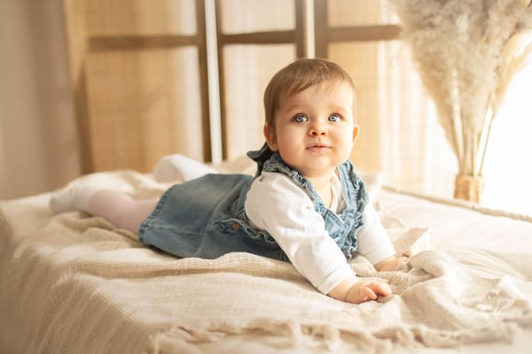 Baby im Jeanskleid liegt auf dem Bauch auf einem hellen Bett und schaut neugierig auf