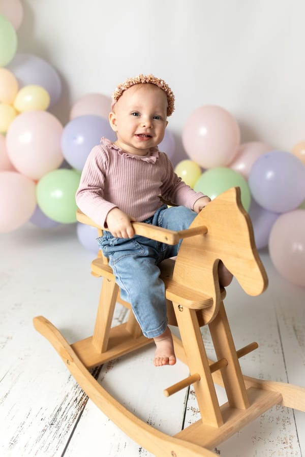 Einjähriges Mädchen sitzt lachend auf einem Holz-Schaukelpferd vor Pastellballons