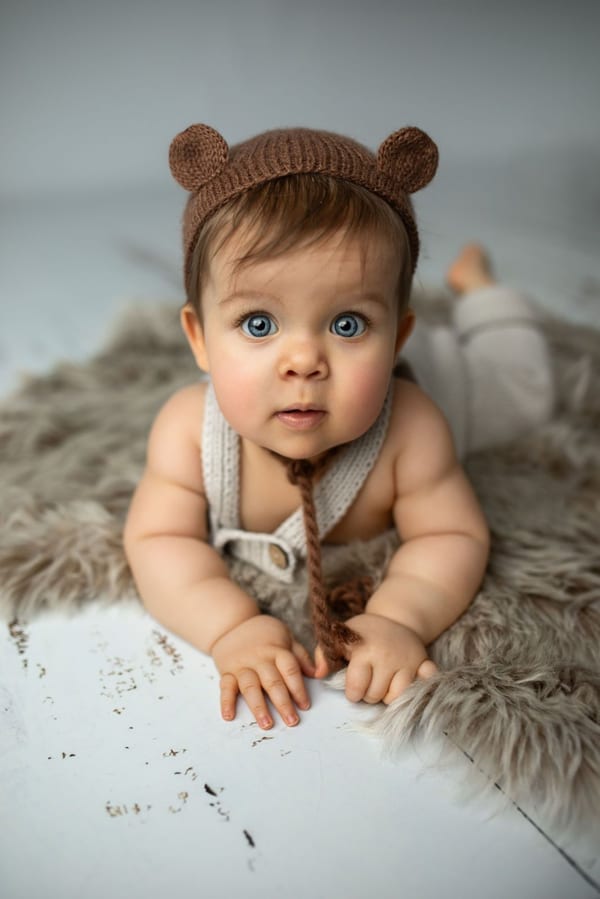 Baby mit gestrickter Bärenmütze liegt auf einem Fell und schaut neugierig in die Kamera