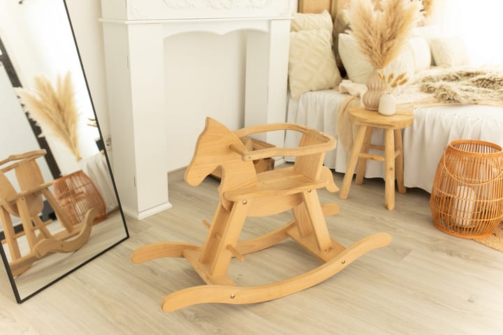 Holz-Schaukelpferd im Atelier vor weißem Kaminsims, daneben Bett mit hellen Kissen, Holzhocker mit Pampasgras und Rattan-Laterne