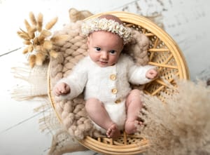 Baby im weißen Strickanzug mit Blütenkranz liegt wach im Rattankörbchen zwischen Pampasgras