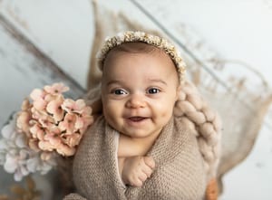 Lachendes Baby mit Blütenhaarband, eingewickelt in eine Strickdecke, daneben rosa Hortensien