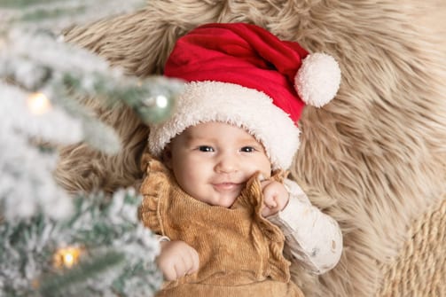 Baby mit roter Weihnachtsmütze liegt lächelnd auf einem hellen Fell, daneben ein verschneiter Tannenzweig