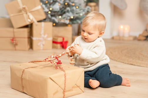 Kleiner Junge im Strickpulli zieht am Geschenkband eines Pakets, dahinter Lichterbaum und Geschenke