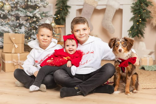 Zwei große Brüder mit ihrer kleinen Schwester im roten Kleid und ihrem Australian Shepherd vor dem Weihnachtsbaum