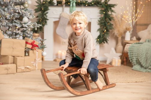 Junge im Rentier-Pulli sitzt auf einem Holzschlitten vor Geschenken und Tannenbaum