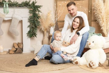 Eltern sitzen mit ihrem Baby auf dem Teppich vor dem weihnachtlichen Kamin, daneben ein großer Teddy