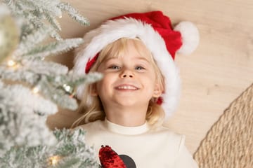 Mädchen mit Weihnachtsmütze liegt lachend auf dem Boden neben einem verschneiten Tannenbaum