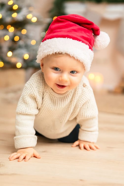 Baby mit roter Weihnachtsmütze krabbelt lächelnd vor dem leuchtenden Tannenbaum