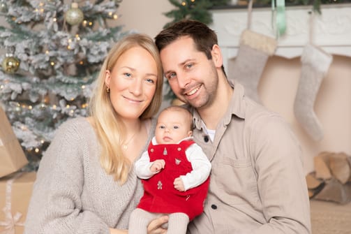 Junge Eltern mit ihrem Baby im roten Weihnachtsstrampler vor dem geschmückten Tannenbaum