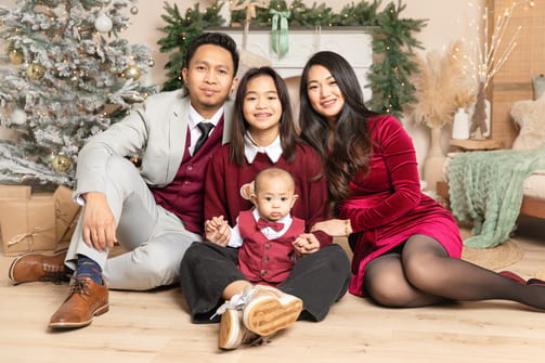 Familie in festlicher Kleidung sitzt mit Tochter und Baby auf dem Boden vor dem Tannenbaum
