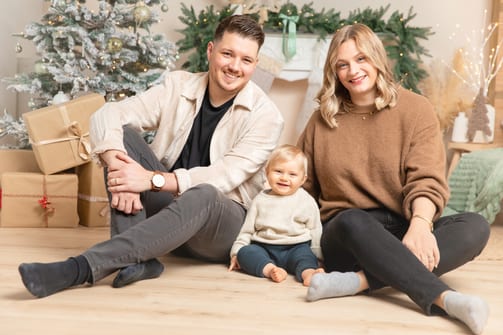 Eltern sitzen lachend auf dem Boden, ihr Baby sitzt zwischen ihnen vor der Tannengirlande