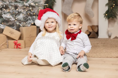 Mädchen mit Weihnachtsmütze und kleiner Bruder mit roter Fliege sitzen vor Tannenbaum und Geschenken