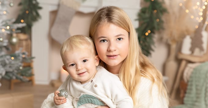 Große Schwester hält ihr kleines Geschwisterchen im Arm vor dem weihnachtlichen Kamin