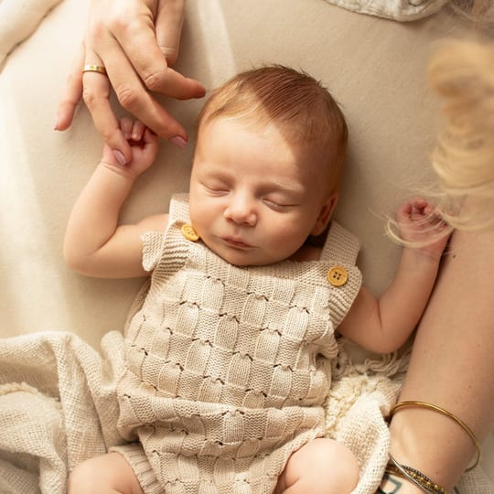 Schlafendes Neugeborenes im Strickstrampler hält einen Finger der Mutter